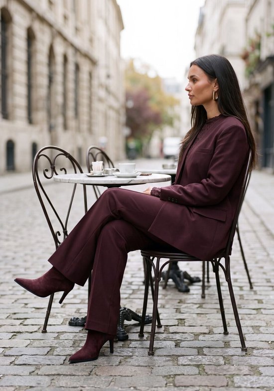 Vrouw in een bordeauxrode broek en colbert, zittend aan een tafel, draagt bordeauxrode suède enkellaarzen met naaldhak.