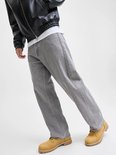 Man in wijde grijze jeans, een zwarte jas en gele laarzen, staand op een lichte achtergrond.