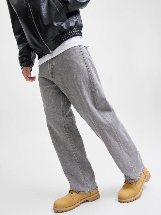 Man in wijde grijze jeans, een zwarte jas en gele laarzen, staand op een lichte achtergrond.