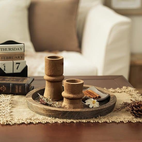 Rond donkerbruin houten dienblad met twee houten kandelaars, kaneelstokjes en een dennenappel op een jute onderzetter.