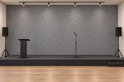 Podium met grijze viltachterwand, sprekerskatheder, microfoonstandaard en luidsprekers.