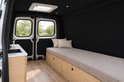 Binnenkant van een camperbusje met zwarte viltwanden, een houten bedframe met beige kussen en een keukengedeelte.