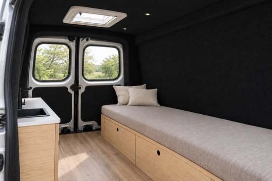 Binnenkant van een camperbusje met zwarte viltwanden, een houten bedframe met beige kussen en een keukengedeelte.