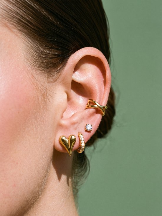 Gouden huggie oorbellen met zirkonia steentjes, hartvormige knopjes en een gekruiste earcuff op een groen achtergrond.