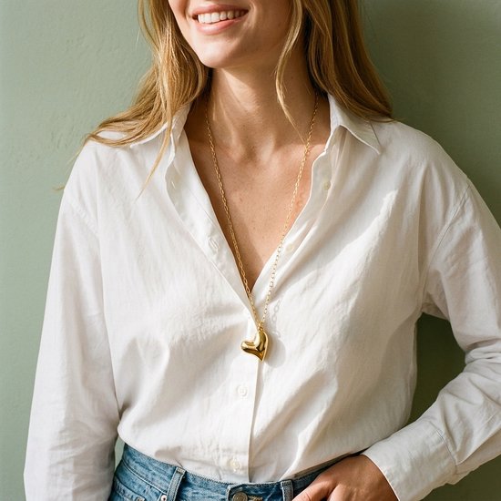 Model draagt een lange goudkleurige ketting met hartvormige hanger over een wit overhemd en blauwe jeans.