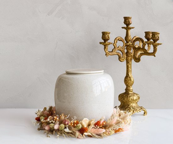 Witte keramische urn met subtiele spikkels, omringd door een droogbloemenkrans, met een gouden kandelaar op de achtergrond.