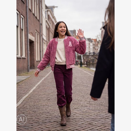Meisje met lang donker haar lacht en wuift in een roze pluizig baseball vest met een witte trui, paarse corduroy broek en bruine laarzen.
