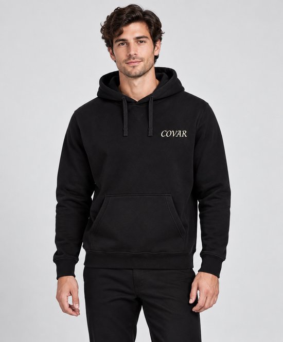 Model draagt een zwarte hoodie met COVAR-logo en zwarte jeans, staand tegen een neutrale achtergrond.