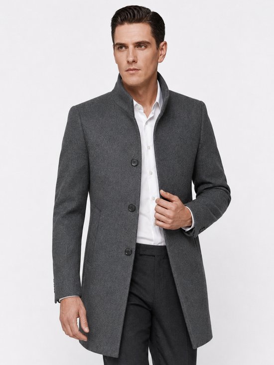 Man met donker haar in een grijze wollen jas met staande kraag, drie knopen, witte blouse, hand in de zak.