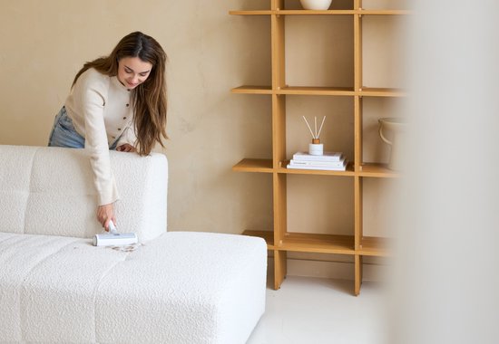 Femme portant un gilet beige et un jean utilise un rouleau anti-peluche sur un canapé blanc.