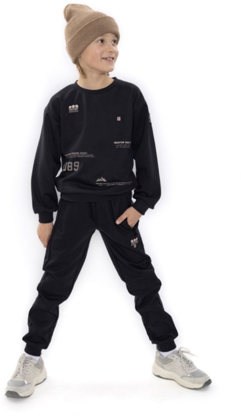 Jongen in een zwarte joggingbroek en sweater, met een bruine muts en grijze sneakers.