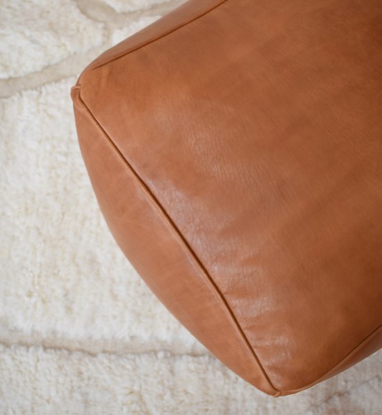 Caramelbruine vierkante poef van leer, op een wit vloerkleed.