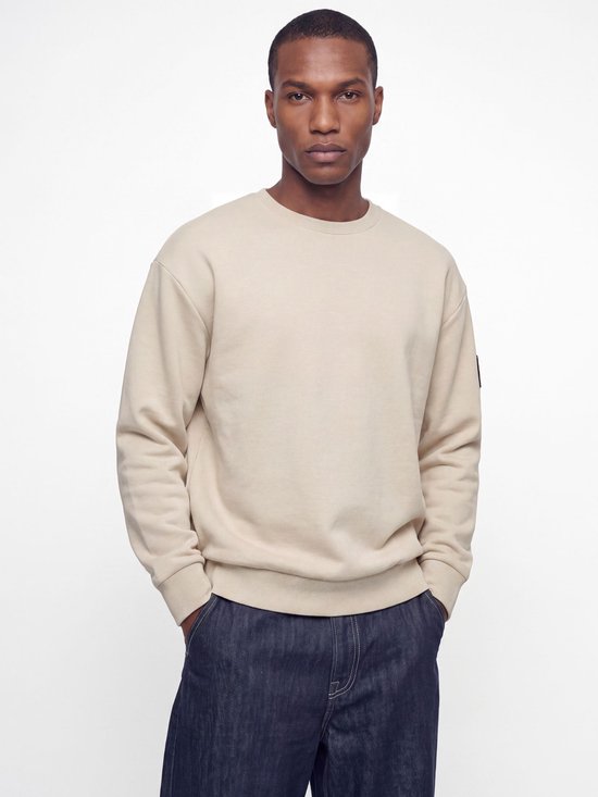 Man in een crème kleurige sweater met ronde hals en donkerblauwe jeans. De sweater heeft lange mouwen en boorden.