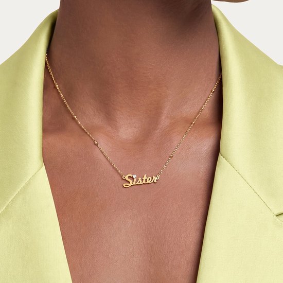 Close-up van een goudkleurige ketting met een 'Sister'-hanger en zirkonia, gedragen bij een limoengroene blazer.