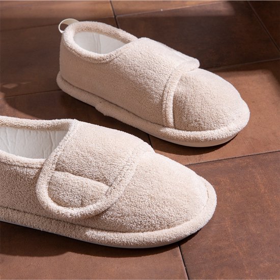 Paar beige pluche pantoffels met een zachte textuur en klittenbandsluiting op een bruine tegelvloer.