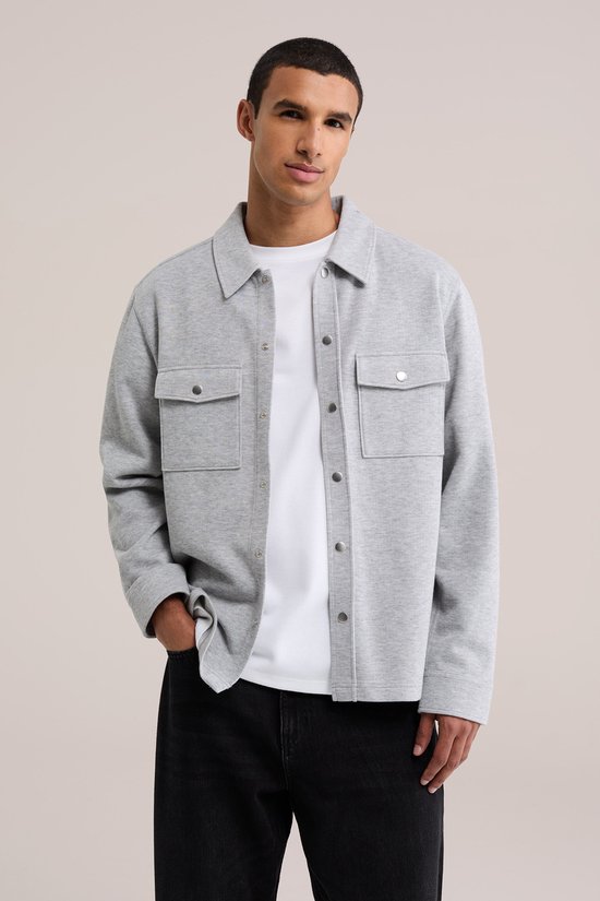 Model draagt een grijs fijngebreid overshirt over een wit T-shirt, met zwarte broek en hand in de zak.