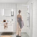Vrouw met bruine handdoek in badkamer, kijkend naar witte douchepaneel.