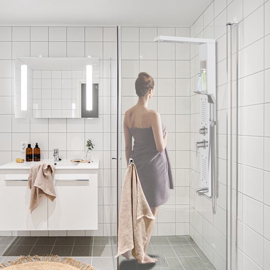 Vrouw met bruine handdoek in badkamer, kijkend naar witte douchepaneel.