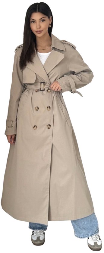 Vrouw draagt lange beige trenchcoat, knoopsluiting, ceintuur, omgeslagen kraag. Witte top, lichtblauwe jeans, sneakers.