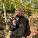 Een lachende vrouw met blonde haren die een beige Phomo camera vasthoudt in een bos.