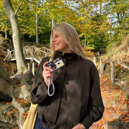 Een lachende vrouw met blonde haren die een beige Phomo camera vasthoudt in een bos.