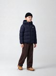 Jongen met donkerblauwe, gewatteerde jas met capuchon en Studio Outdoors logo, draagt zwarte muts en bruine broek.