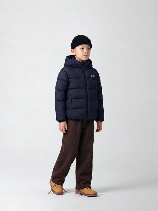 Jongen met donkerblauwe, gewatteerde jas met capuchon en Studio Outdoors logo, draagt zwarte muts en bruine broek.
