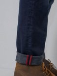 Detail van de omgeslagen onderkant van een donkerblauwe jeans met rode bies en witte stiksels.