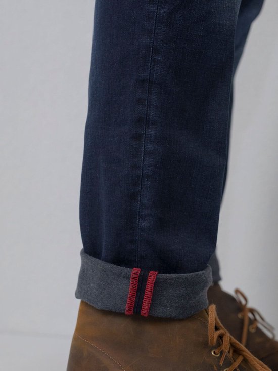Detail van de omgeslagen onderkant van een donkerblauwe jeans met rode bies en witte stiksels.