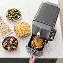 Zwarte Ninja heteluchtfriteuse met een gebraden kip in de lade, naast aardappeltjes, friet en koekjes.