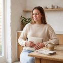 Een lachende vrouw met een beige trui zit aan een houten tafel, met een crème warmtekussen in haar handen. Een kopje en een servet staan naast haar.