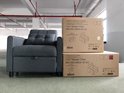 Grijze slaapbank met vierkante knopen op de rugleuning en armleuningen, naast twee kartonnen dozen met '3 in1 Sleeper Chair' erop.