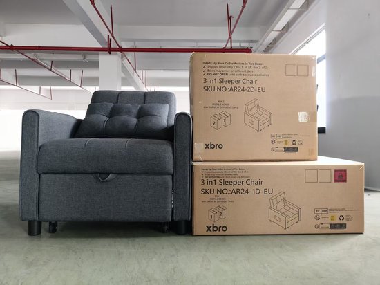 Grijze slaapbank met vierkante knopen op de rugleuning en armleuningen, naast twee kartonnen dozen met '3 in1 Sleeper Chair' erop.