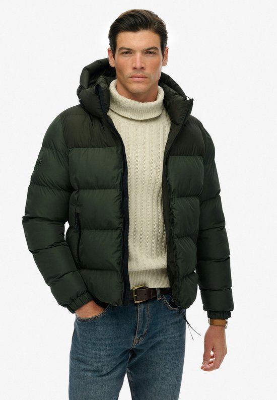 Man met donkergroene, gewatteerde jas met capuchon, beige coltrui en spijkerbroek.