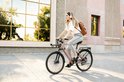 Vrouw met witte top en grijze broek op champagne gouden elektrische fiets met rugzak.