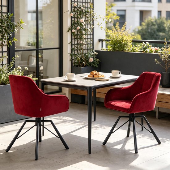 Twee bordeauxrode fluwelen stoelen met zwarte poten aan een tafel op een balkon met planten.