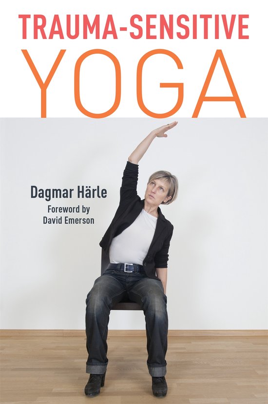 Vrouw met korte blonde haren, gekleed in een wit T-shirt, zwart jasje en jeans, zit op een stoel en strekt haar rechterarm omhoog. "Trauma-sensitive Yoga" staat bovenaan.
