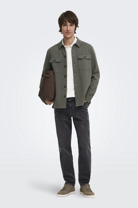 Man in olijfgroene utility blouse, wit poloshirt, grijze jeans, bruine tas en sneakers.