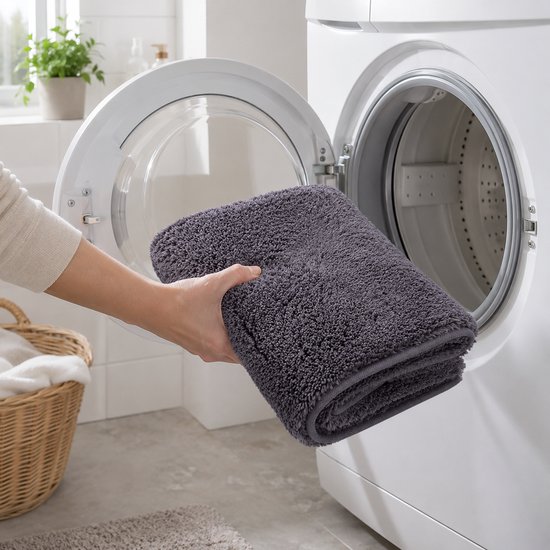 Hand die een donkergrijze, opvouwbare badmat in een open wasmachine plaatst.