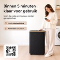 Een vrouw scannt met haar telefoon een QR-code naast een zwarte wasmand met bamboehouten afwerking. De wasmand staat in een wasruimte.