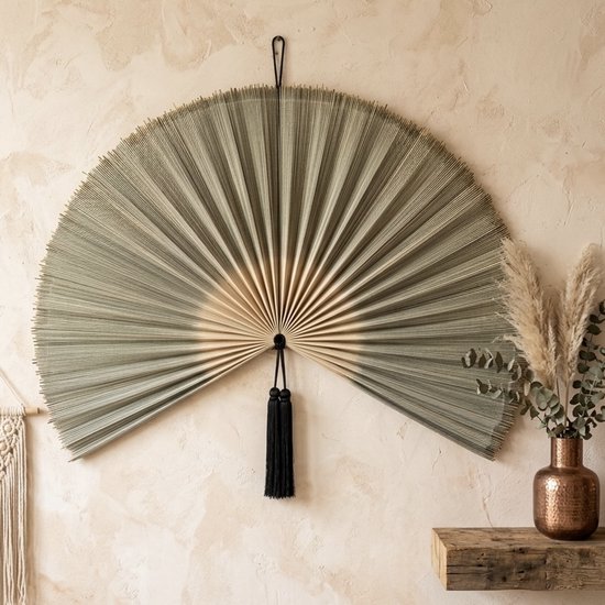 Grote waaier van geweven materiaal in grijs en beige, hangend aan een muur boven een koperen vaas met pampasgras.