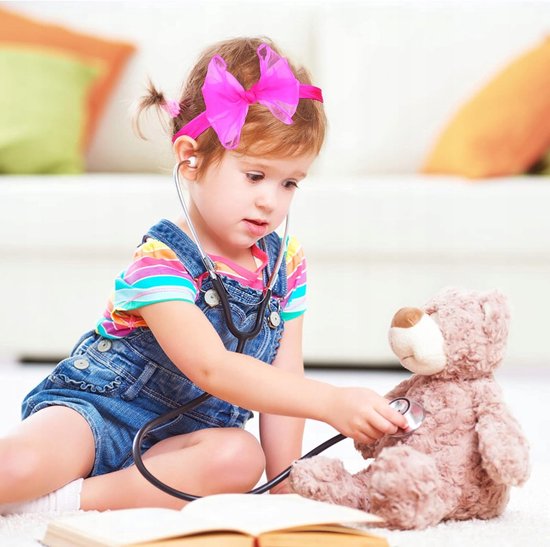 Een jong meisje met een roze strik als haarband, gekleed in een spijkerbroek met bretels en een gestreepte top, luistert naar haar teddybeer met een stethoscoop.