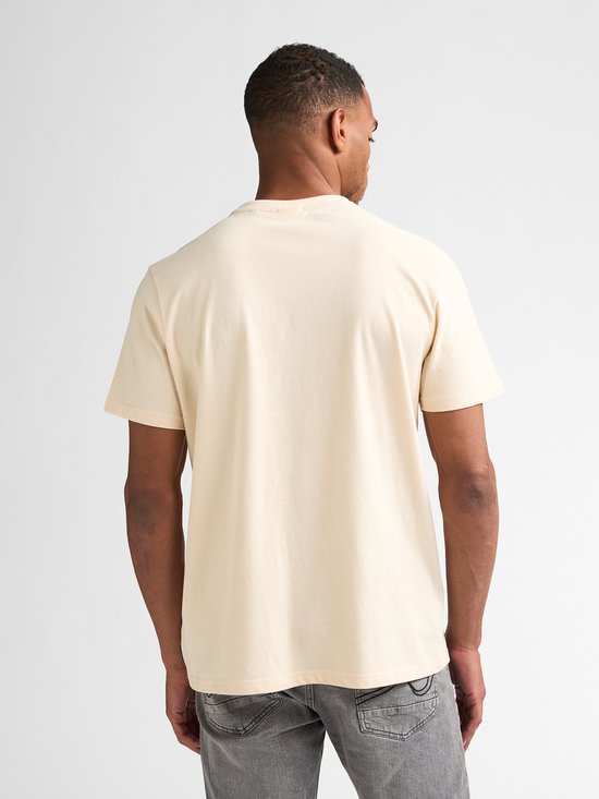 Man staat met de rug naar de camera, draagt een crème T-shirt met korte mouwen en grijze jeans.