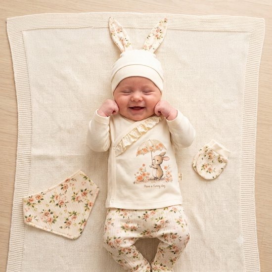 Baby in romper met konijnenprint, bijpassende muts, broek, slab en wanten. Bloemenpatroon in zachte tinten.