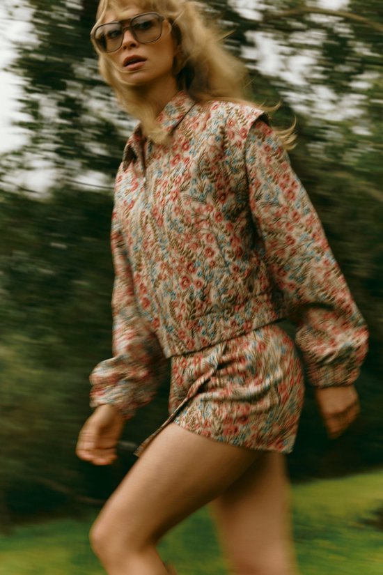 Model in beweging in een gebloemd jacquard jasje en bijpassende korte broek. Zonnebril op.