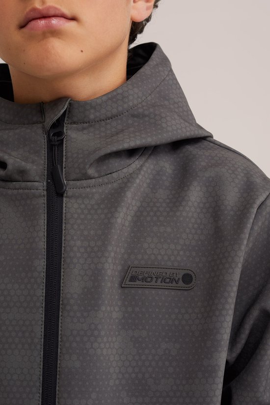 Close-up van een donkergrijze softshell jas met capuchon. De jas heeft een subtiel geometrisch patroon en een logo met tekst 'DEFINED BY MOTION'.