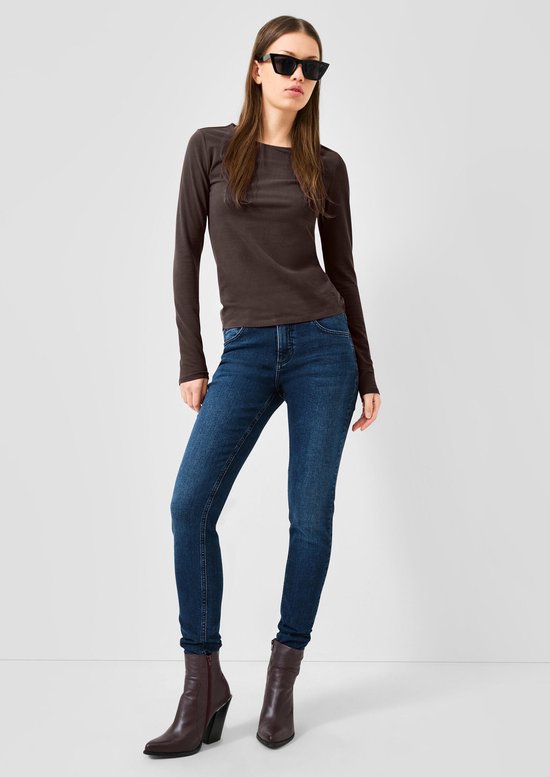 Vrouw draagt donkerbruine longsleeve, zonnebril, blauwe jeans en enkellaarsjes.