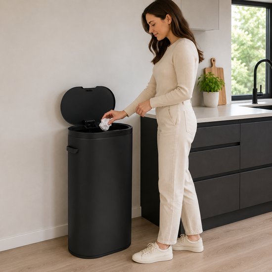 Vrouw met een lichte trui en broek gooit papier in een hoge, zwarte, ovale prullenbak in een keuken.