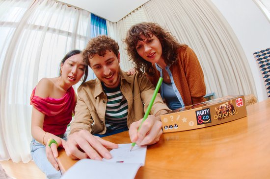 Drie vrienden, twee vrouwen en een man, spelen het spel Party & Co. De man tekent op een papier, de vrouwen kijken mee.