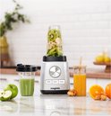 Zilverkleurige Magimix blender met fruit en groenten in de beker, naast een glas groene smoothie en een glas oranje sap.